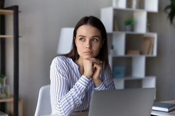 Serious thoughtful employee sitting at table with laptop, looking at window away, thinking over business problems, project, future career vision, feeling doubts, uncertain, dreaming and pondering