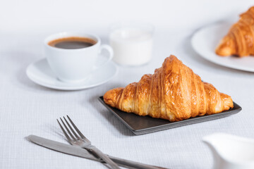 Croissant in bright white environment, with cup of coffee and milk and other croissant in blurry background. Simple and elegant breakfast setting and scene.