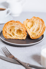 Croissant in bright grey environment and cut to two pieces, with cup of coffee and milk and other croissant in blurry background. Simple and elegant breakfast setting and scene.