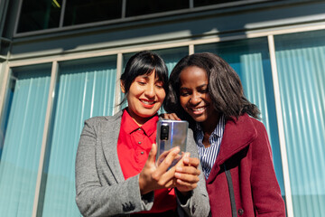 African girl and Latin woman smiling while watching funny multimedia content on their mobile phone.