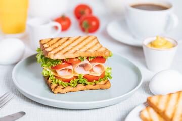Toast sandwich with gouda cheese and turkey ham filled with tomato and lettuce on a bright background with ingredients in blurry background. Served with coffee and orange juice and eggs