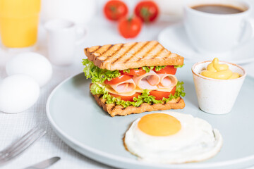 Toast sandwich with gouda cheese and turkey ham filled with tomato and lettuce on a bright background with ingredients in blurry background. Served with coffee and orange juice and eggs