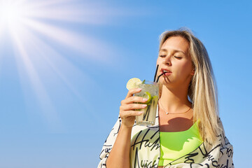 Beautiful woman in a yellow swimsuit is standing in the sun and enjoying a cold mojito in hot...