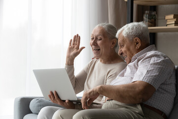 Happy middle aged elderly retired family couple waving hand, looking at laptop screen, starting online conversation using computer video call application, communicating distantly with grown children.