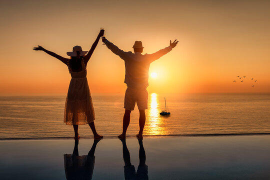 A happy couple on holidays celebrates the summer sunset over the sea by the swimming pool with a glass of wine