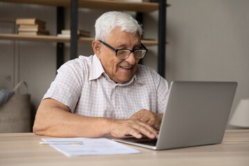 Fototapeta premium Smiling mature elderly retired man in eyeglasses working on computer, sitting at table at home or office, enjoying communicating in social network, checking email, web surfing or shopping online.