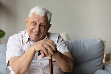 Happy dreamy senior elderly grandfather holding hands on walking wooden cane, resting on sofa....