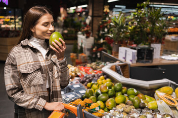Female customer in supermarket smelling mango. Woman chooses fruit in the supermarket - obrazy, fototapety, plakaty
