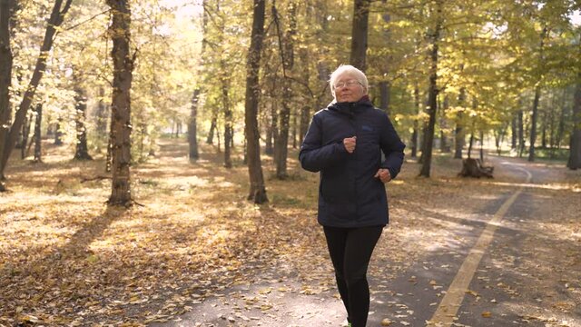 Close-up of aged gray-haired female suffering from heart burn during running in autumn park. Elderly woman jogger feeling pain during active cardio training and getting a heart attack
