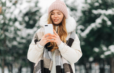 woman in a knitted hat and a warm coat , walks in the winter park and uses a mobile phone