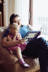 Dad reading bedtime story to his little daughter. They are sitting on big comfy armchair near window