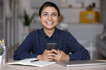Happy millennial Indian ethnic girl chatting online on mobile phone at work table, typing, looking away, posing with toothy smile. Employee woman texting clients in office, consulting internet