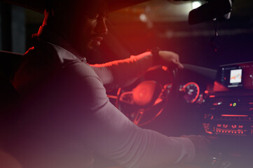 cute bearded strong young man in white shirt sitting in luxury sport muscle car lit with red light in underground garage