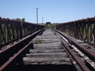 Old abandoned train track passes over an old bridge and continues towards infinity among the vegetation and illuminated by the sun. Metal structure of iron and wood worn by the passage of time.