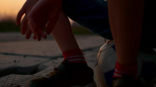 Close-up of a guy sitting on a soccer ball. A teenager in sneakers sits on a ball resting after the game, in the rays of the setting sun. Youth and boyish ball games.
