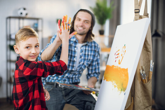 Happy cute boy and handsome man smiling and looking at camera while giving high five to each other. Father and son using easel for drawing at home.