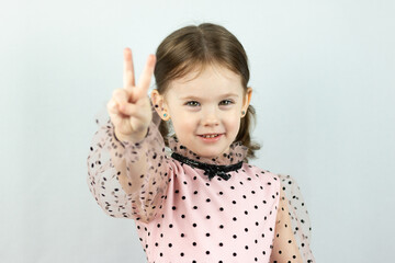Smiling little girl with two ponytails in a dress with polka dots on a white background shows two fingers raising her hand forward. Studio photo