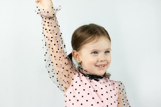 A smiling little girl with two ponytails in a dress with polka dots on a white background looks to the side raising her hand up. Studio photo