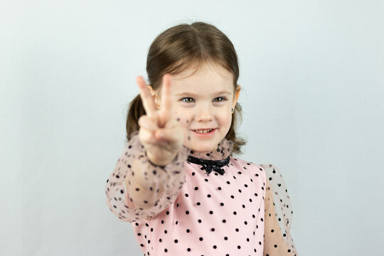Smiling little girl with two ponytails in a dress with polka dots on a white background shows two fingers raising her hand forward. Studio photo