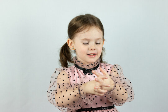 Smiling little girl with two ponytails in a dress with polka dots on a white background holds both hands in front of her. Studio photo