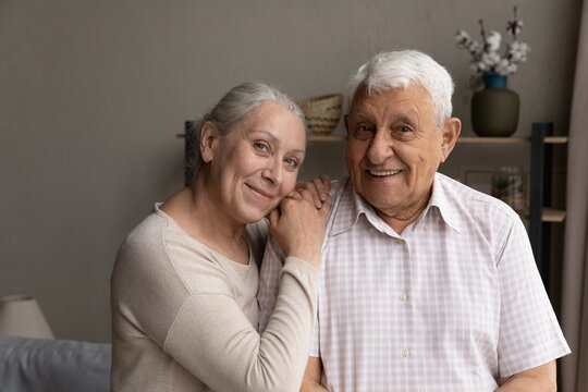 Portrait Of Smiling Sincere Affectionate Older 60s Beautiful Hoary Woman Putting Head On Shoulder Of Happy Loving Elderly Retired 80s Husband, Candid Joyful Mature Family Couple Looking At Camera.