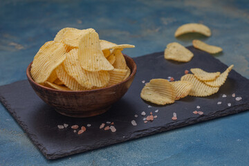 Crispy potato chips in a wooden bowl on blue background. Salty crisps scattered around.