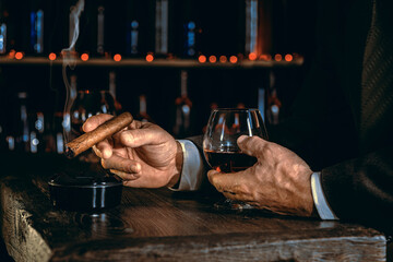Man's hands with a cigar, elegant glass of brandy on the bar counter. Alcoholic drinks, cognac, whiskey, port, brandy, rum, scotch, bourbon. Vintage wooden table in a pub at night