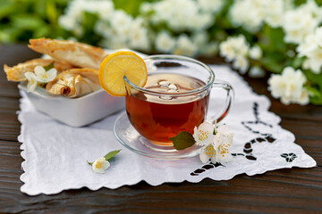 Glass cup with tea and petals, lemon, croissants, embroidered napkin, jasmine branches on a wooden table. Outdoor, picnic, brunch. Selective focus with short DOF.