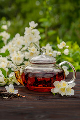 Steamed glass teapot with jasmine tea among flowering bushes. Outdoor, picnic, brunch. Floral background in blur.