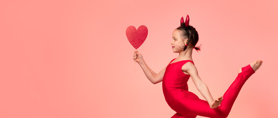 Little smiling girl with pigtails in costume of little devil holding red heart in hand and jumping against red background