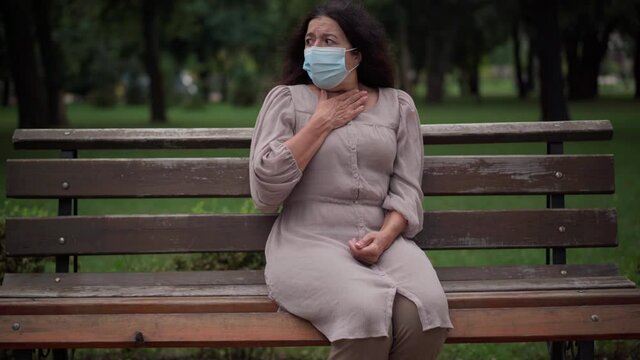 Front view portrait stressed Caucasian senior woman in Covid-19 face mask coughing sitting on bench in park outdoors. Worried frightened retiree with symptoms of coronavirus illness looking around