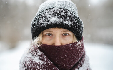 Blonde woman in beanie hat and nose covered in scarf looks at camera in winter snowfall. Nordic...