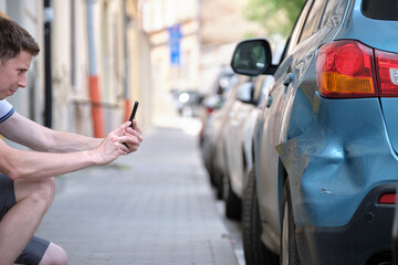 Stressed driver taking picture on sellphone camera of smashed vehicle parked on street side calling for emergency service help after car accident. Road safety and insurance concept