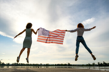 Fototapeta na wymiar Two young friends women with USA national flag jumping up together outdoors on lake shore. Patriotic girls celebrating United States independence day.