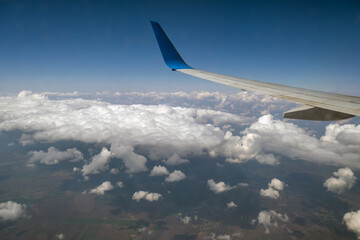 View of jet airplane wing from inside flying over white puffy clouds in blue sky. Travel and air transportation concept