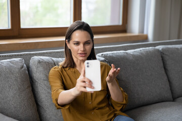 Beautiful millennial telephone caller woman talking on mobile phone, making conference call from home couch, holding smartphone, speaking, gesturing. Blogger recording video. Communication