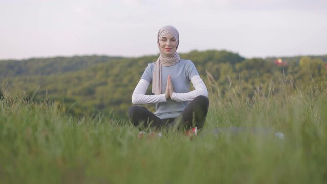 Peaceful young lady in hijab meditating with closed eyes and namaste gesture in the park. Pretty Muslim woman sitting on couch in yoga pose, finding inner balance