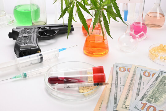Photo contains pistol, test tubes, colored fluids in chemical retorts and flasks, natural cannabis leaves, blood tests, inoculation syringes. 100 dollar banknotes on a white lab table. Laboratory