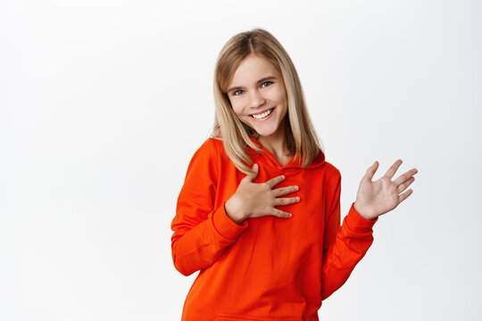 Image of cute little blond girl saying hi, waving hand and smiling friendly, introducing herself, greeting, standing in red casual hoodie over white background