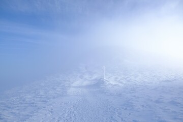 Fototapeta premium A frozen pole marking trail in the mountains by path on foggy and sunny winter day. Babia Gora peak in background. Diablak, Beskid Zywiecki, Poland