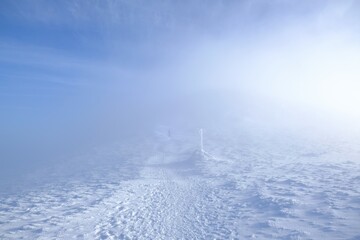 A frozen pole marking  trail in the mountains by  path on foggy and sunny winter day. Babia Gora peak in background. Diablak, Beskid Zywiecki, Poland