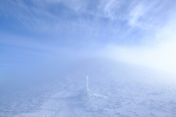 A frozen pole marking  trail in the mountains by  path on foggy and sunny winter day. Babia Gora peak in background. Diablak, Beskid Zywiecki, Poland