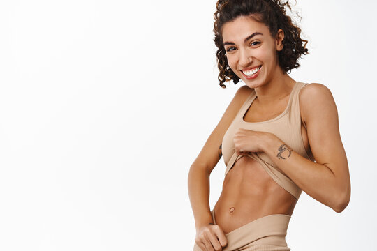 Smiling fitness girl in sportsbra looking at her abs, abdominal muscles with pleased face, effect after workout in gym, standing over white background