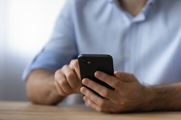 Crop close up man holding smartphone, sitting at wooden desk, chatting in social network, typing on screen, scrolling, surfing internet, browsing apps, focus on modern mobile device in male hands