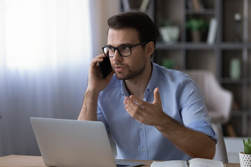 Confident businessman in glasses talking on phone, sitting at work desk with laptop, serious focused man negotiating with business partner or colleague, consulting client, holding interview