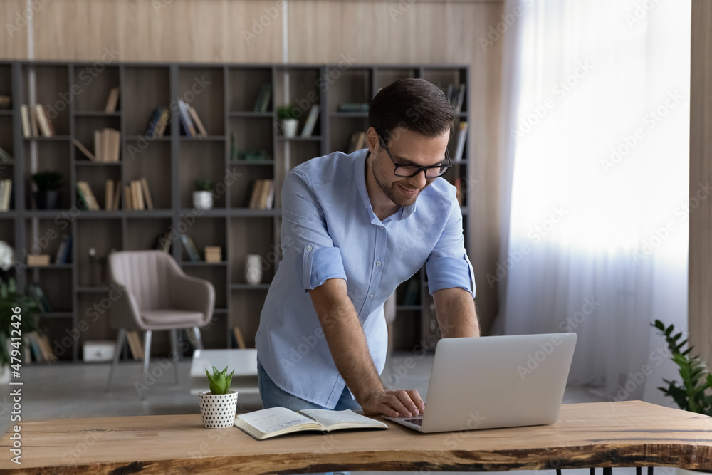 Poster Smiling businessman in glasses using laptop, standing at work desk, confident satisfied man freelancer or student looking at computer screen, typing writing message business email, home office