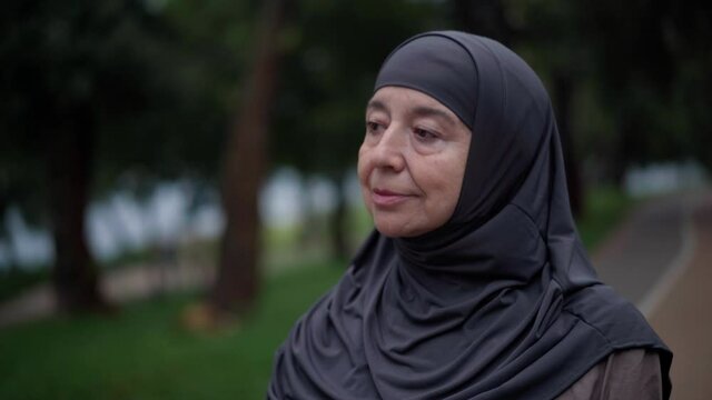 Close-up portrait of confident gorgeous Muslim senior woman in hijab strolling outdoors looking away. Beautiful Middle Eastern retiree walking in spring summer park enjoying resting. Leisure concept