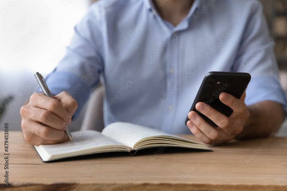 Poster Cropped close up businessman holding smartphone, writing down important information, taking notes, sitting at wooden desk, entrepreneur planning workday schedule, student watching webinar, studying