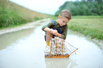 A boy, 7 years old, is playing with a small sailing ship in a puddle after the rain.
