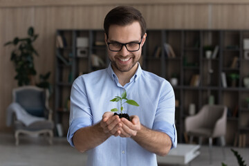 Smiling businessman wearing glasses holding small green plant sprout with soil, standing, happy entrepreneur employee with growing tree, startup project, profit, investment and growth concept - Powered by Adobe